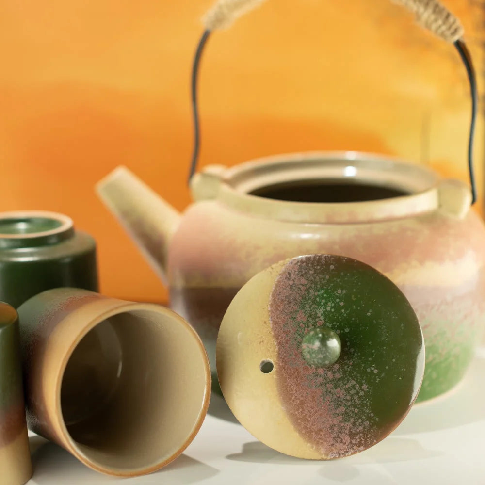 A ceramic teapot set with a speckled green, beige, and brown glaze, including a teapot with a woven handle, a small matching cup, and a lid shown open in the foreground against a warm orange background.