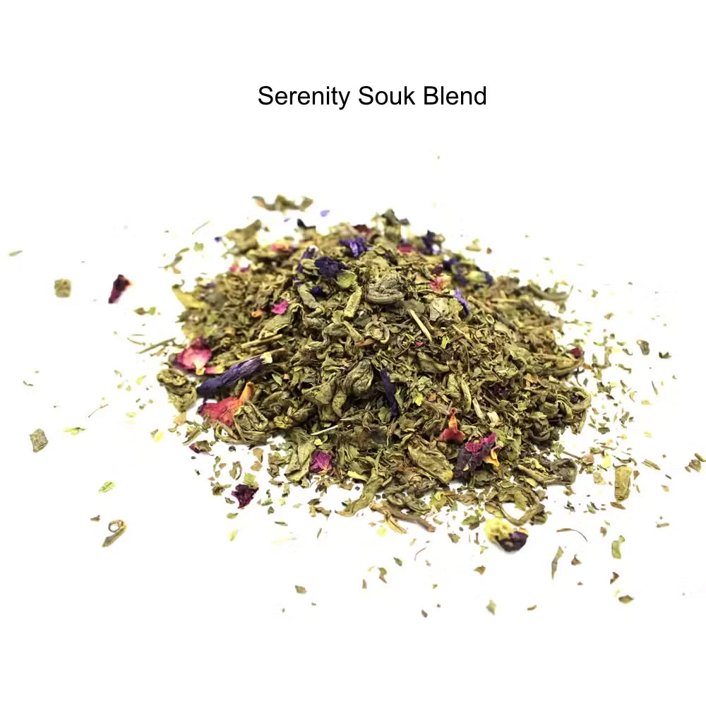 A small pile of loose herbal tea blend labeled 'Serenity Souk Blend' on a plain white background. The blend is primarily green with visible dried flower petals in pink, red, and purple tones.