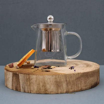 Clear glass teapot with a stainless steel infuser and lid, displayed on a round wooden slice against a dark gray background.
