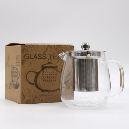 Glass teapot with metal lid and handle next to its packaging box on a white background