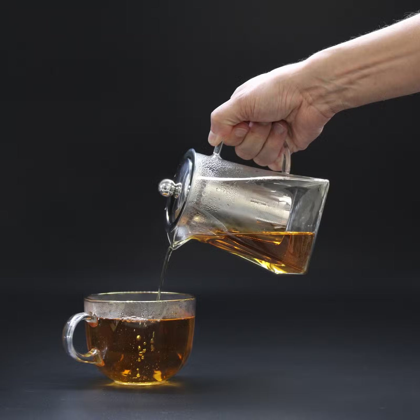 Hand pouring tea from a glass teapot into a clear glass mug on a dark background