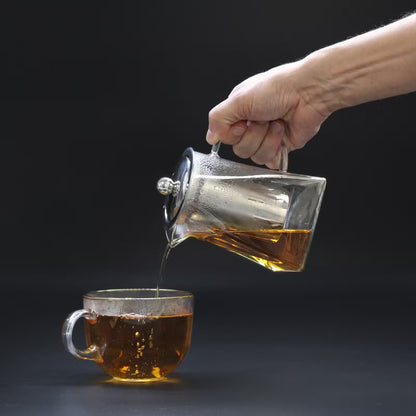Hand pouring tea from a glass teapot into a clear glass mug on a dark background