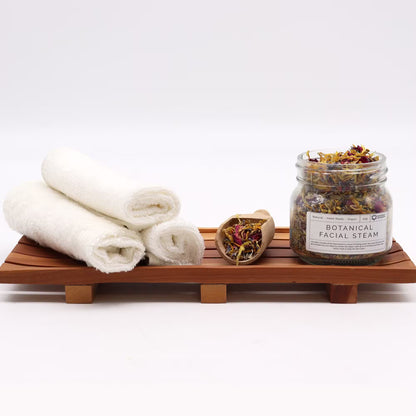 Jar of botanical facial steam with towels and a wooden tray on a white background