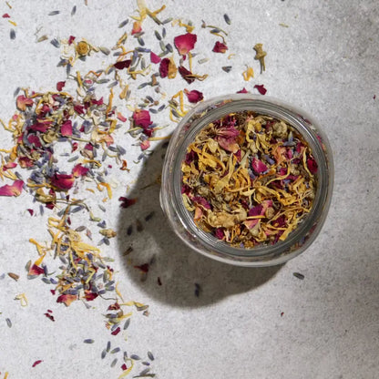 An open clear glass jar filled with dried flower petals and loose botanicals, photographed from above on a light textured surface with scattered petals around it.