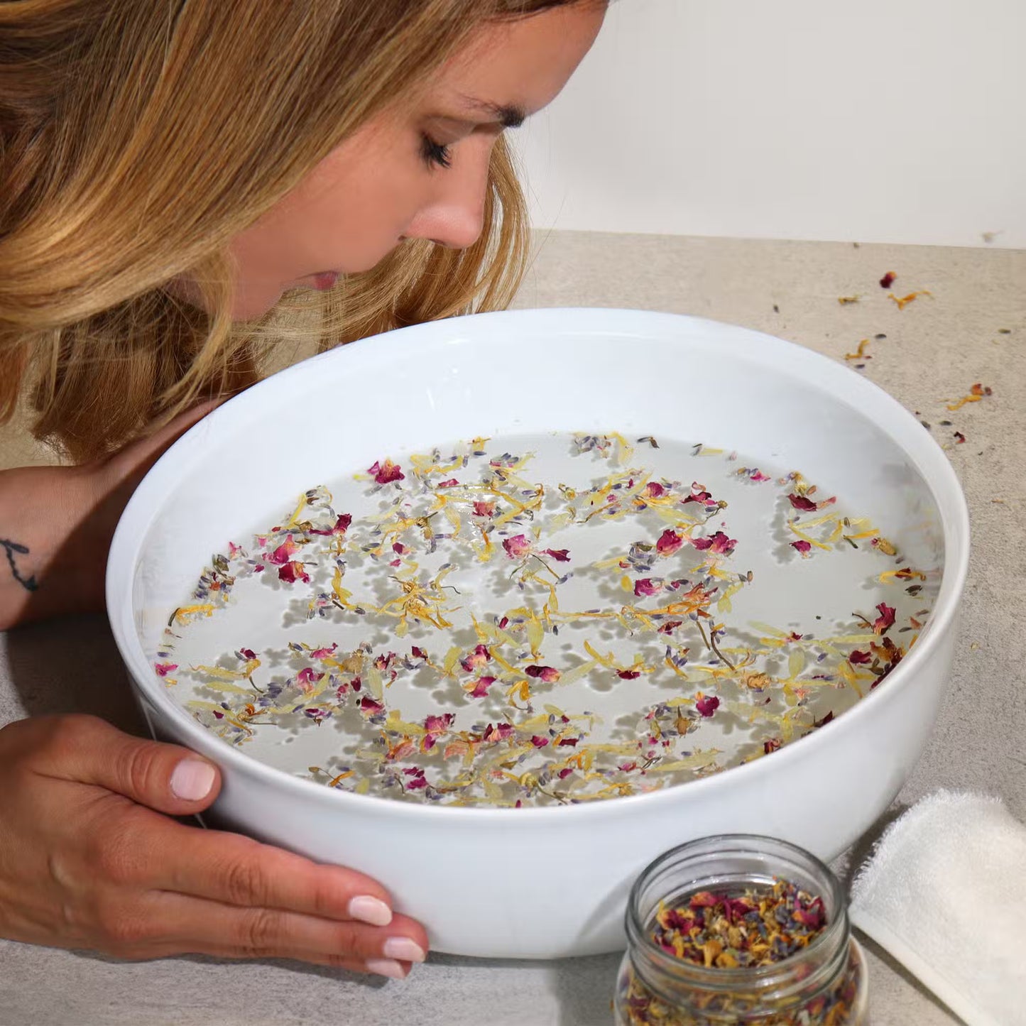 Woman smelling a bowl of water with flowers, next to a jar of flowers on a towel.