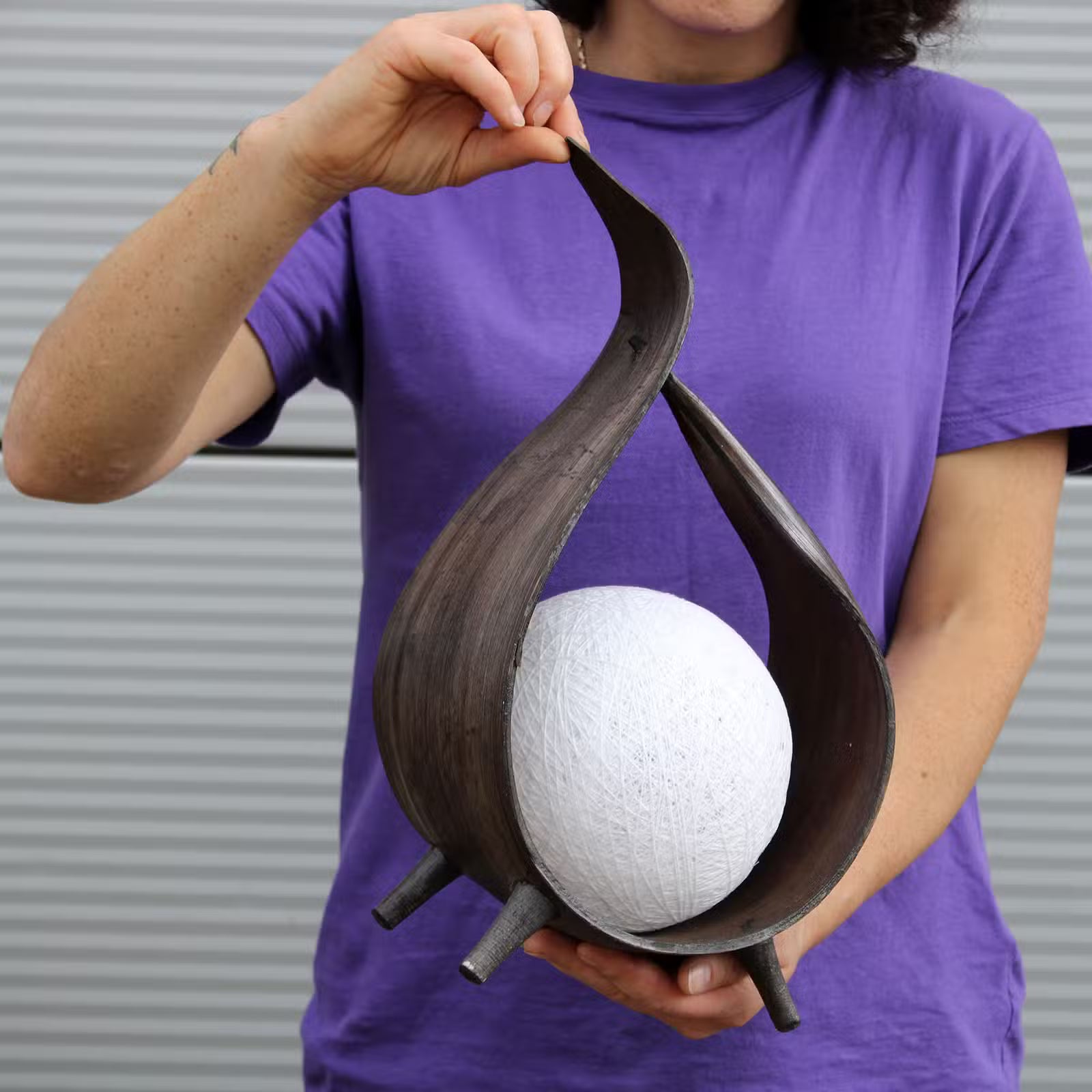 A person in a purple t-shirt holds a dark brown sculptural wooden bowl with a white textured sphere inside, set against a gray corrugated metal background.