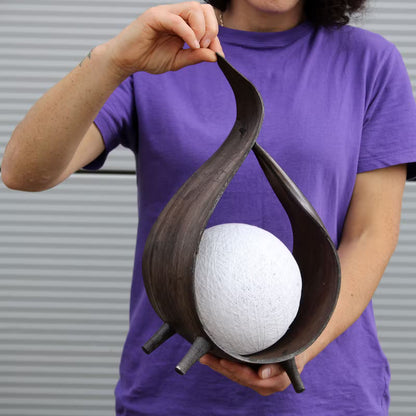 A person in a purple t-shirt holds a dark brown sculptural wooden bowl with a white textured sphere inside, set against a gray corrugated metal background.