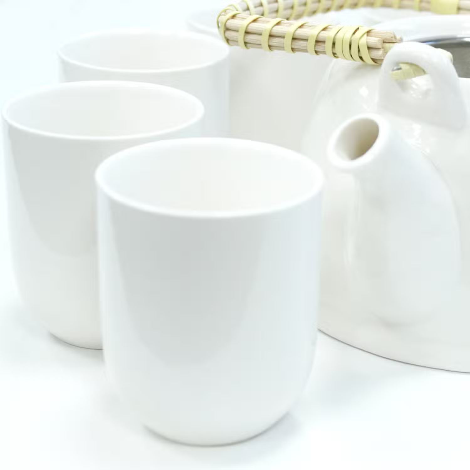 Three glossy white ceramic cups arranged in front of a matching white teapot with a light natural bamboo handle on a clean white background.