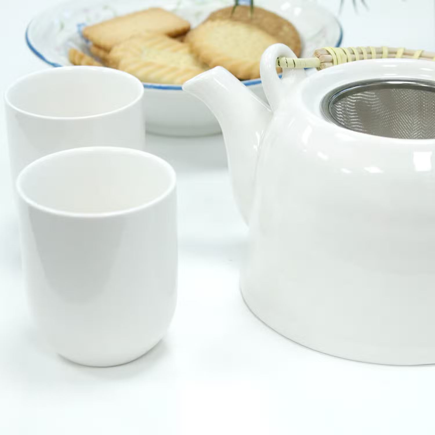 A white ceramic teapot with a metal tea infuser inside, shown alongside two matching white ceramic cups on a white surface. A blurred plate of cookies appears in the background.