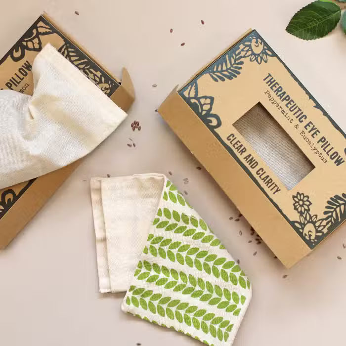 Two kraft paper boxes for herbal eye pillows are arranged on a light beige surface, with one box showing a folded off-white cloth pillow and the other showing a white cloth pillow with a green leaf pattern. Text on the boxes reads 'Therapeutic Eye Pillow' and 'Clear and Clarity'.