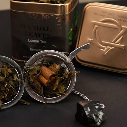 Tea leaves in a strainer with a tea canister and packaging on a dark surface