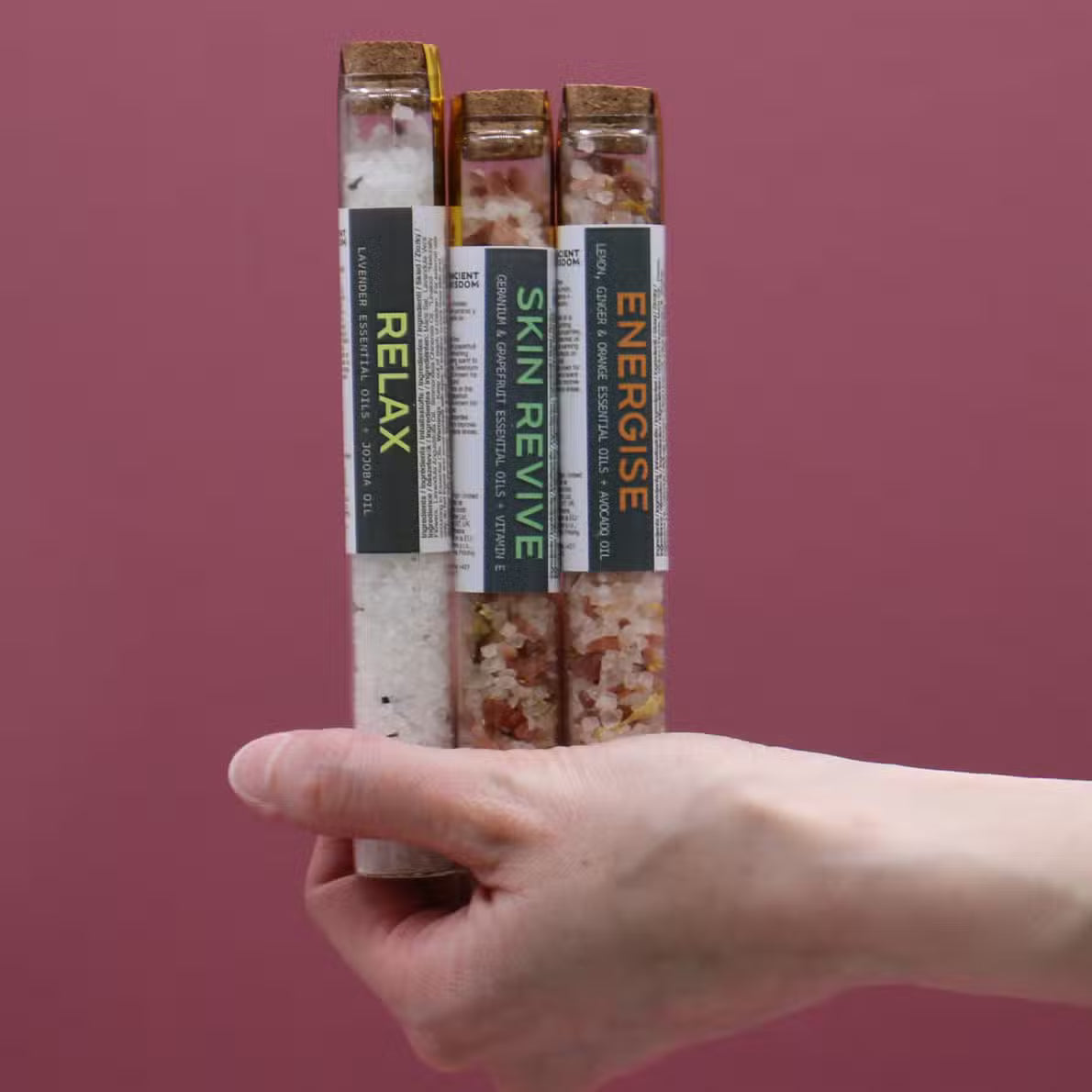Hand holding three bath salt sticks with labels on a pink background