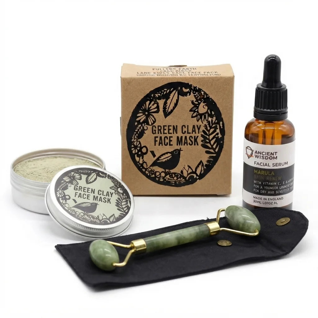 A facial care gift set on a white background, including a Green Clay Face Mask tin and box, an amber dropper bottle of Ancient Wisdom Facial Serum, a green jade roller, and a black velvet pouch.
