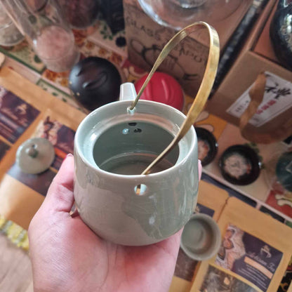 Hand holding a green ceramic mug with a gold spoon, against a blurred background of a table with various items.