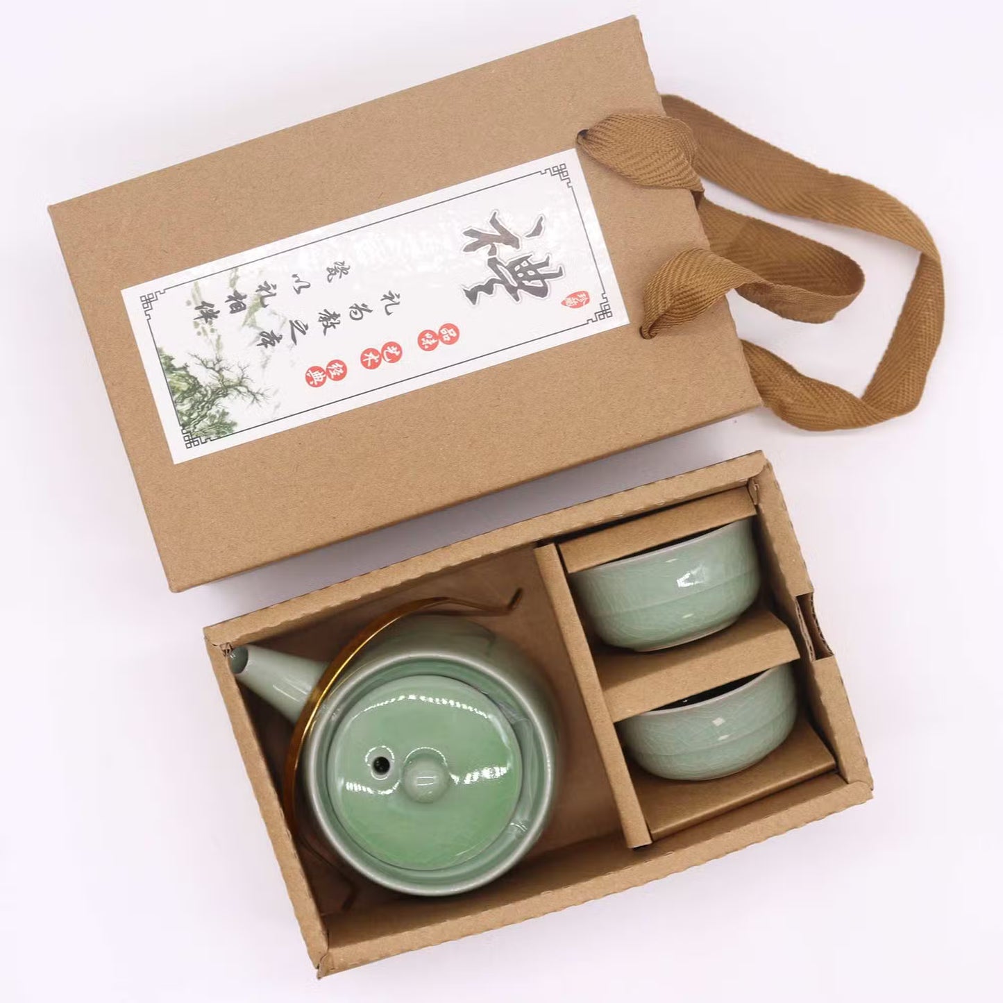A ceramic tea set in a light green glaze, including a teapot and two small cups, presented in a brown cardboard gift box with a fabric handle and a printed label.
