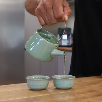 Person pouring tea from a green teapot into two cups on a wooden surface.