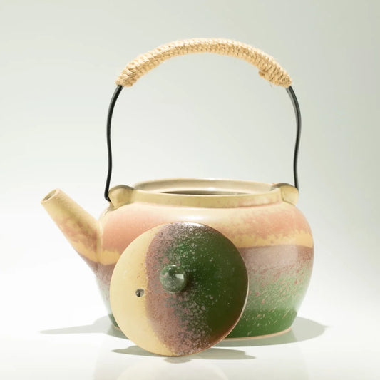 A ceramic teapot with a rounded body, a matching lid, and a woven straw handle, shown on a clean white background. The teapot features a marbled glaze with beige, pink, and green colors, and the lid has a green and brown pattern.