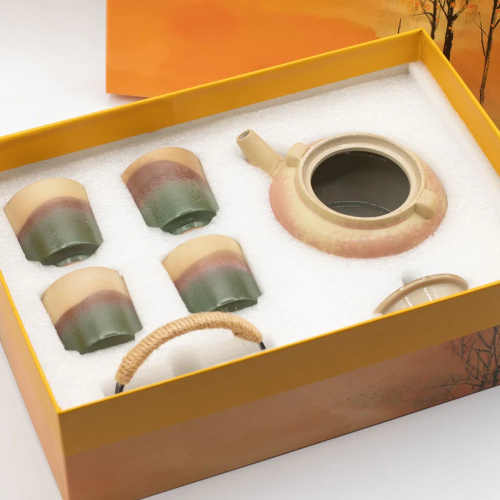 A ceramic tea set displayed inside a yellow gift box with white foam insert. The set includes a teapot with a beige and peach glaze, four matching small cups with beige, brown, and green gradients, and a small woven carrying handle.