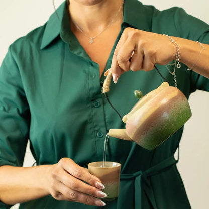 A person in a dark green collared shirt pours tea from a ceramic teapot with a brown and green gradient glaze into a matching small cup. The person is wearing silver jewelry, and the background is a clean, neutral off-white wall.