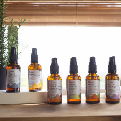 Row of six bottles of body oil on a wooden surface with a window and plant in the background.