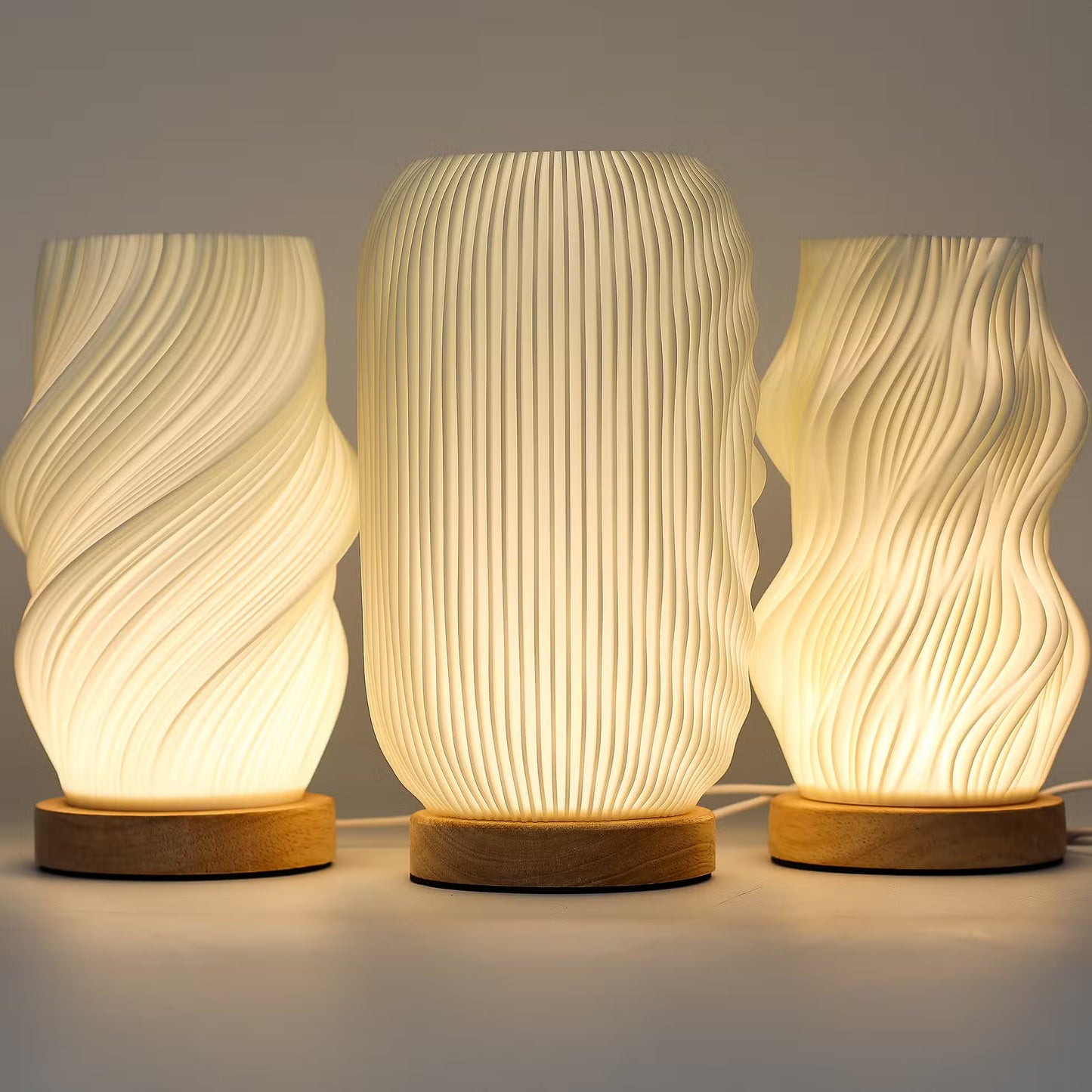 Three modern table lamps with glowing warm white ribbed and wavy translucent shades on round natural wood bases, arranged against a neutral gray background.