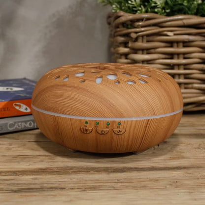 A wooden diffuser with a warm honey-toned wood grain finish sits on a rustic wooden table. The diffuser features a rounded, egg-shaped body with a white LED light band around its middle and three small control buttons on the front.
