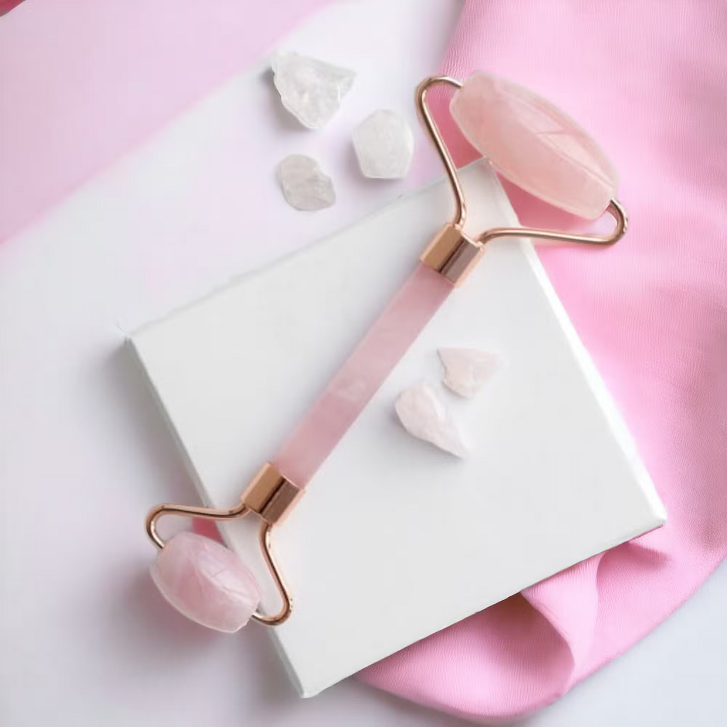 A rose pink jade roller with rose gold metal handles and polished pink stone ends, displayed diagonally on a white box with pink and white fabric background and small crystal props.