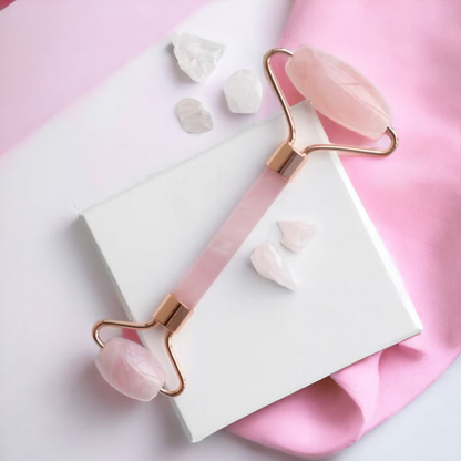 A rose pink jade roller with rose gold metal handles and polished pink stone ends, displayed diagonally on a white box with pink and white fabric background and small crystal props.