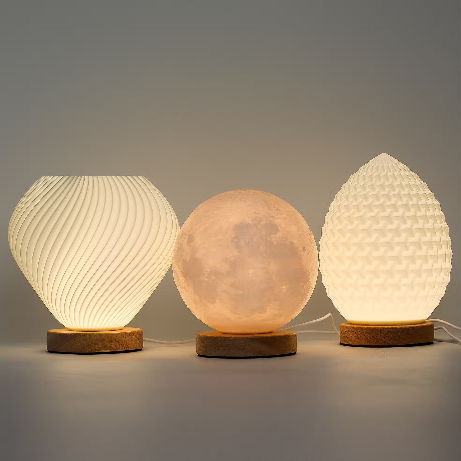 Three illuminated table lamps in different shapes sit on wooden bases against a plain gray studio background. The left lamp is ribbed and rounded, the center lamp is a glowing moon sphere, and the right lamp resembles a pinecone with a textured surface.