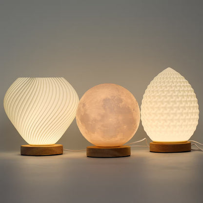 Three illuminated table lamps in different shapes sit on wooden bases against a plain gray studio background. The left lamp is ribbed and rounded, the center lamp is a glowing moon sphere, and the right lamp resembles a pinecone with a textured surface.