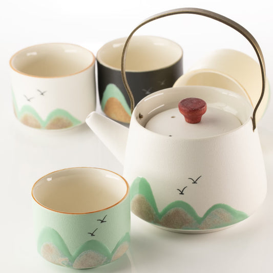 A ceramic tea set with a teapot and cups decorated with a minimalist landscape pattern of green hills, beige rocks, and small black birds. The teapot has a dark metal handle and a reddish-brown lid knob, while the cups have a glossy finish.