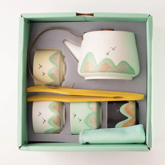 A ceramic tea set gift set in a mint green box, featuring a white teapot with green and beige mountain designs, two matching cups, a yellow wooden spoon, a folded mint green cloth, and a small black-and-white item, all arranged on a gray insert.