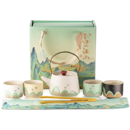 A ceramic tea set displayed in front of its illustrated gift box on a white background. The set includes a white teapot with a green mountain-patterned base, four matching cups in white, mint green, and dark gray, yellow chopsticks, and a decorative mat with a matching scenic print.