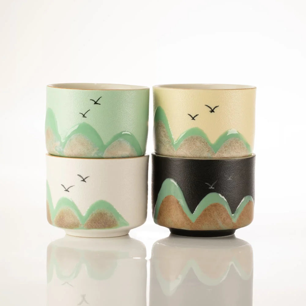 Four stacked ceramic cups or small bowls with a mountain-and-birds motif, shown on a clean white background with a soft reflection below. The set includes pastel green, cream, white, and dark charcoal/black designs with tan mountain shapes and small black birds.