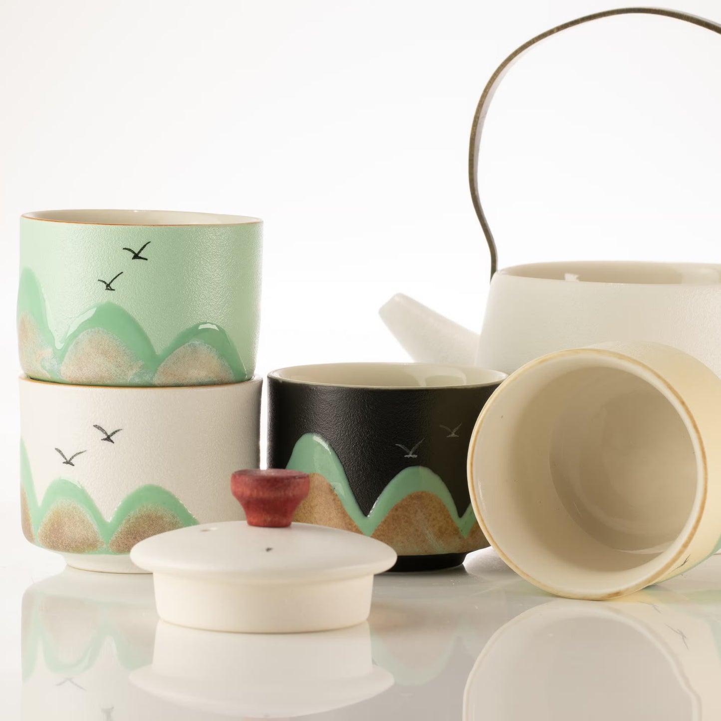 A ceramic tea set with multiple cups and a teapot on a reflective white surface. The set includes a stack of cups decorated with green and beige mountain patterns and small black birds, a black cup with a matching green and beige pattern, a white teapot with a metal handle, and a white lid with a small red knob.