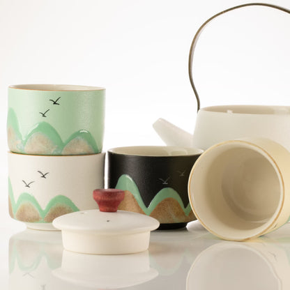 A ceramic tea set with multiple cups and a teapot on a reflective white surface. The set includes a stack of cups decorated with green and beige mountain patterns and small black birds, a black cup with a matching green and beige pattern, a white teapot with a metal handle, and a white lid with a small red knob.