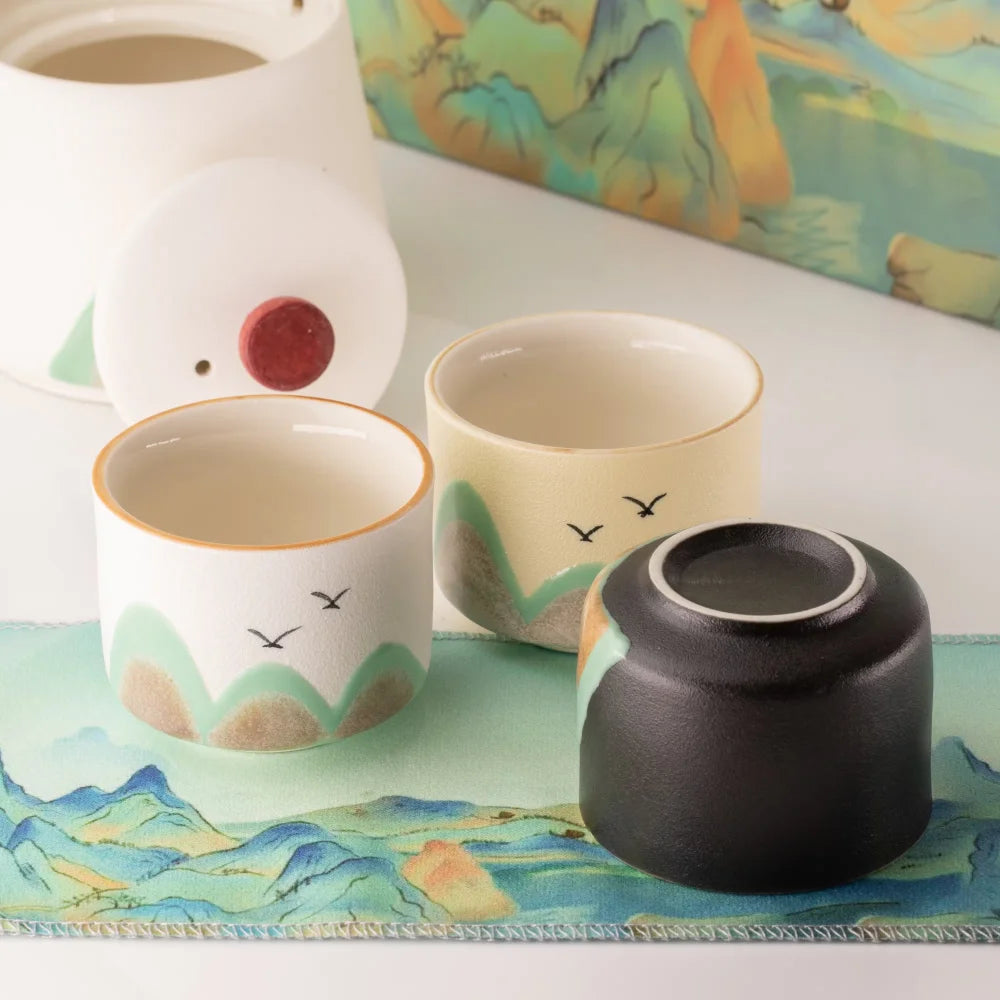 Three ceramic tea cups with a minimalist landscape design are arranged on a matching scenic mat. One cup is shown upside down with its lid, revealing a small red button.