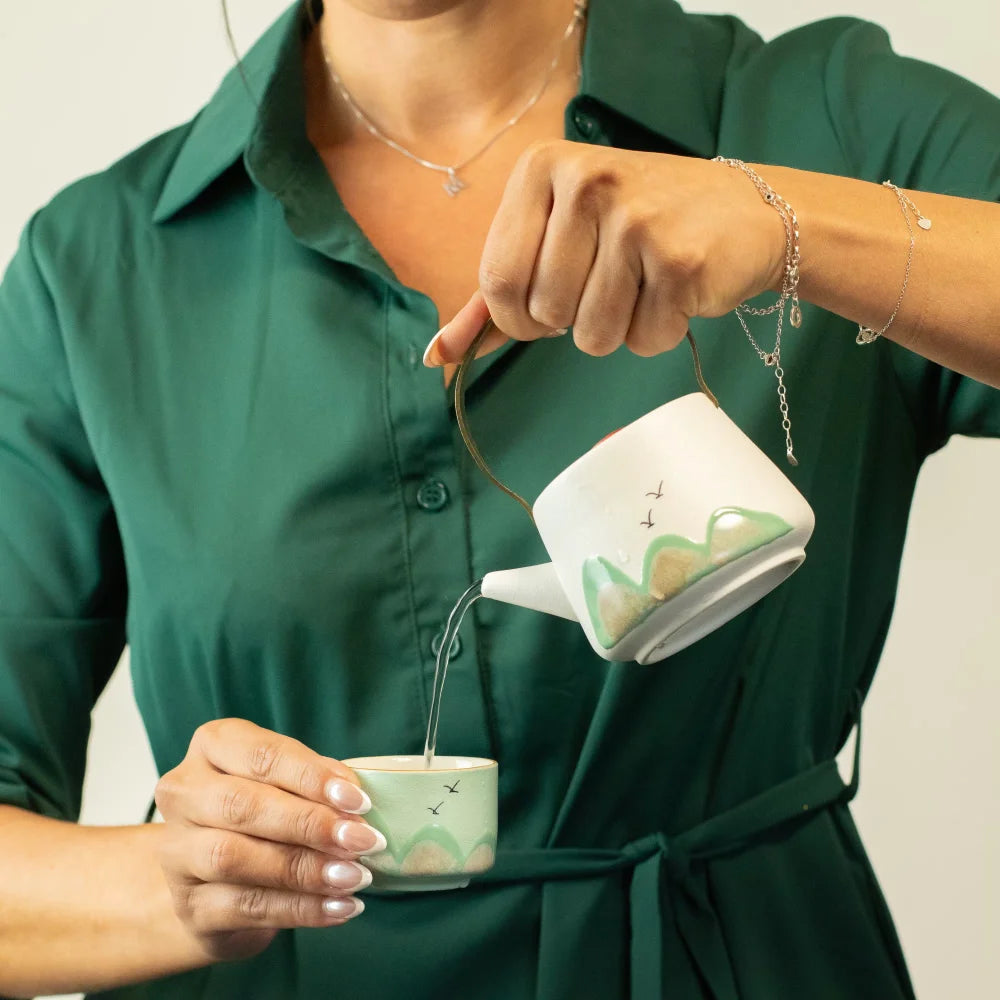 A person in a dark green shirt pours hot water from a white ceramic teapot with a green landscape design into a matching small cup. The teapot and cup have delicate hand-painted green and gold details, and the person is wearing silver jewelry.