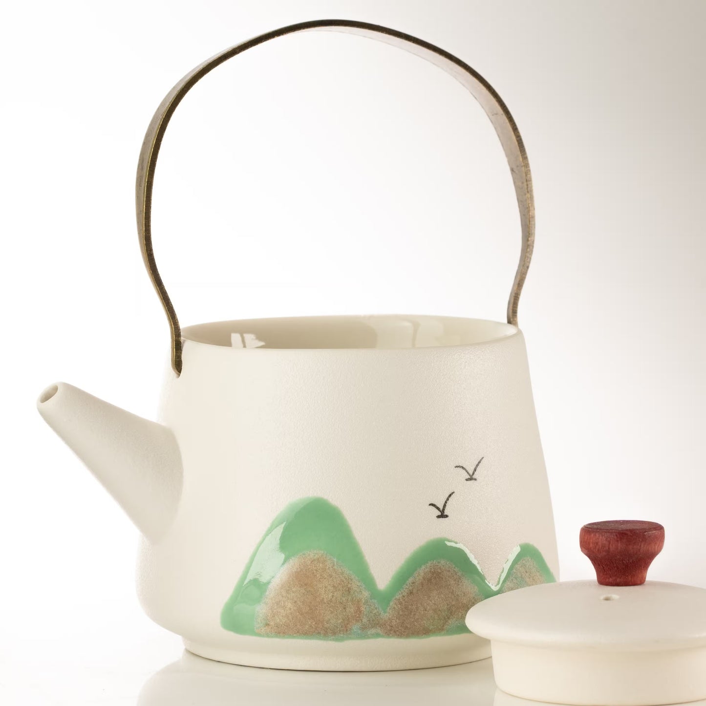 A white ceramic teapot with a metallic handle, decorated with a green and beige mountain landscape design on the front. A matching lid with a reddish-brown wooden knob is shown next to it on a clean white background.