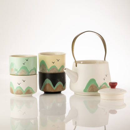 A ceramic tea set on a reflective white surface, featuring a white teapot with a dark metal handle, a matching white lid, two stacked cups in mint green and black-and-cream mountain designs, and a small cup in pale green and cream.