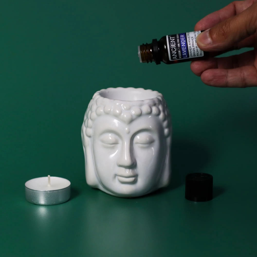 White Buddha head oil burner with a hand holding a bottle of essential oil against a green background