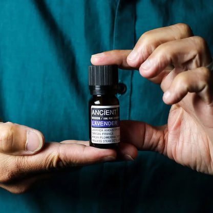 A person holding a small amber glass bottle of Ancient Lavender essential oil over their fingers, with a dark teal shirt in the background.