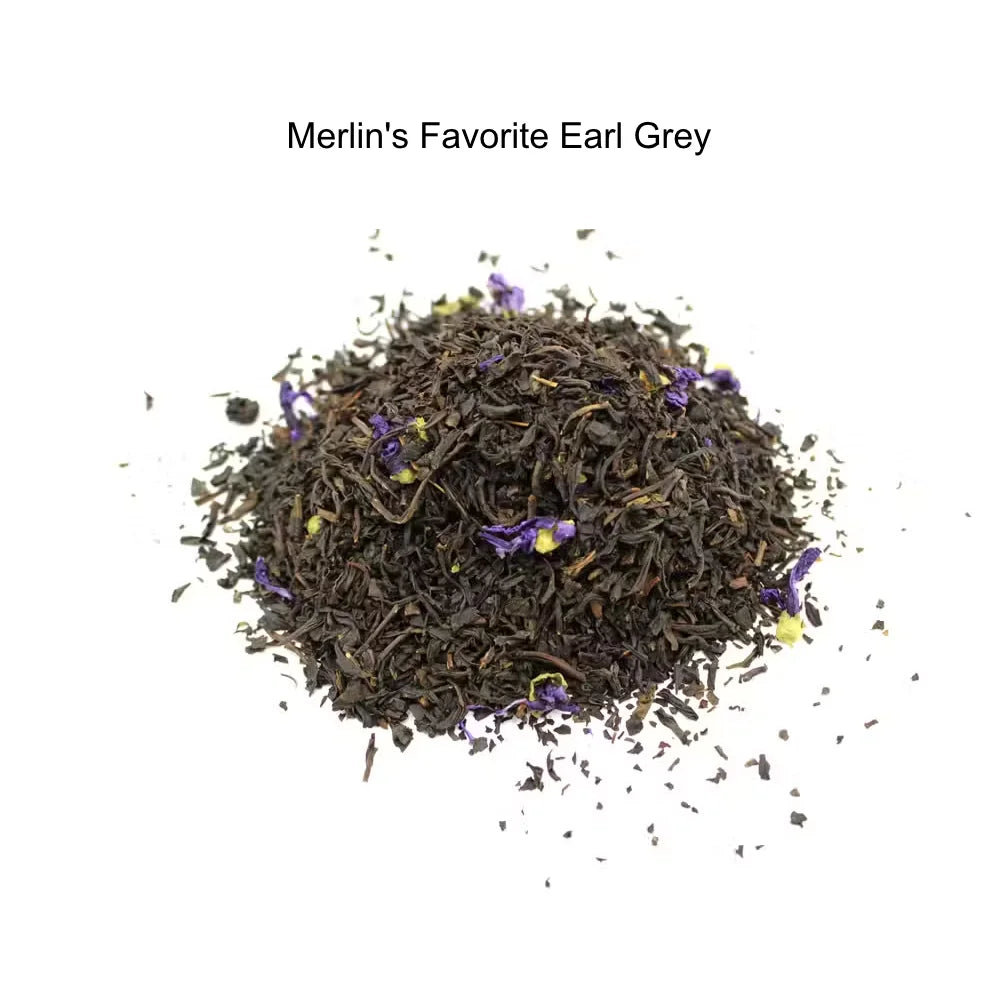 A small mound of loose-leaf tea labeled 'Merlin's Favorite Earl Grey' on a plain white background. The tea is predominantly very dark brown to black with scattered small purple flower petals mixed in.