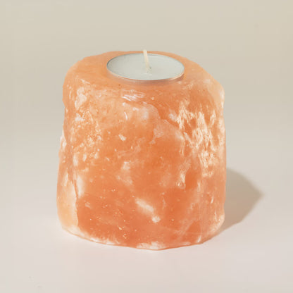 A natural pinkish-orange salt candle holder made from a rough salt crystal, featuring a circular cutout at the top holding a small white tea light candle, set against a plain light beige background.