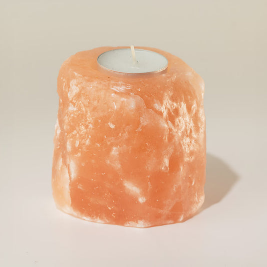 A natural pinkish-orange salt candle holder made from a rough salt crystal, featuring a circular cutout at the top holding a small white tea light candle, set against a plain light beige background.