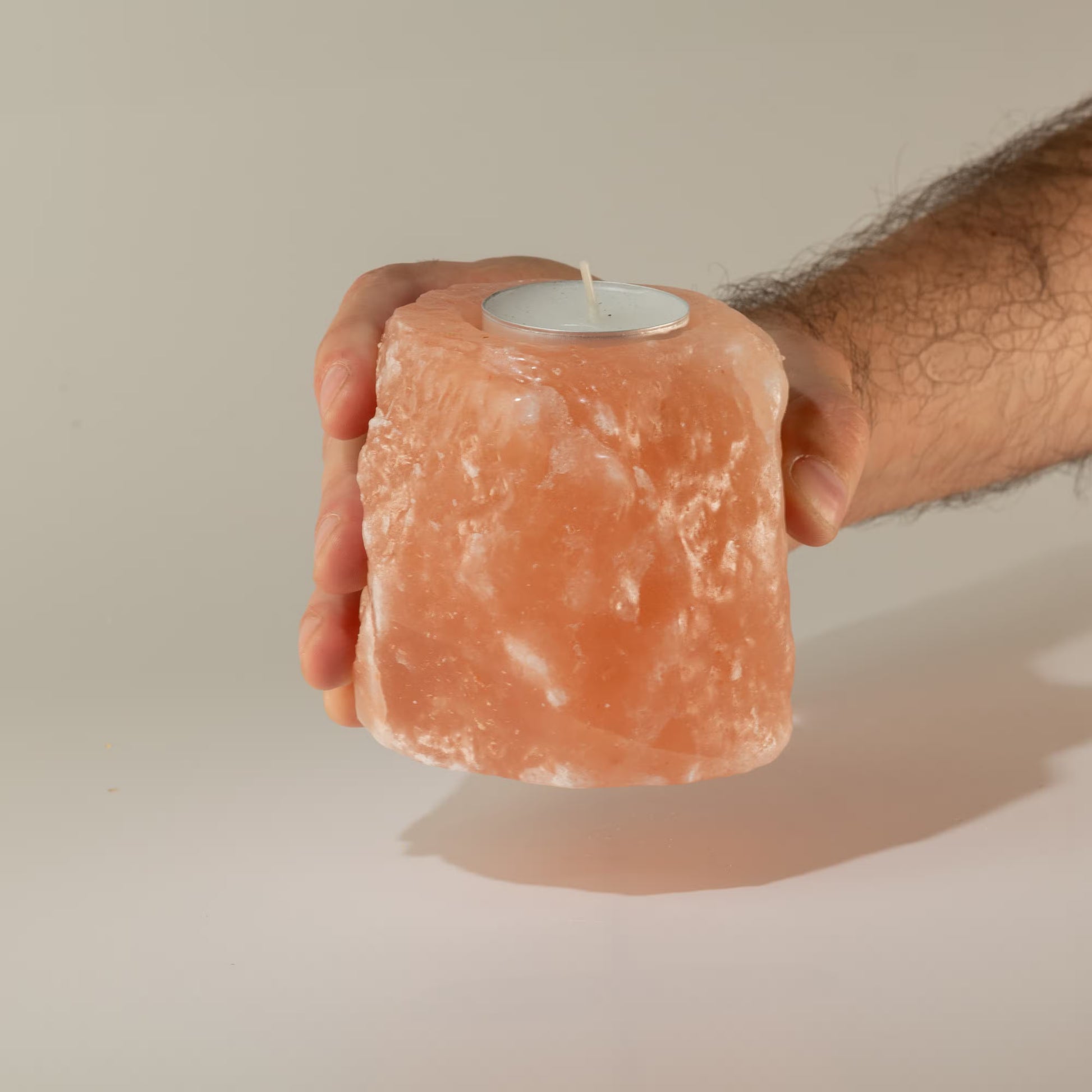 A hand holding a pink salt stone candle holder with a small white tea light candle inserted at the top, set against a plain light gray studio background.