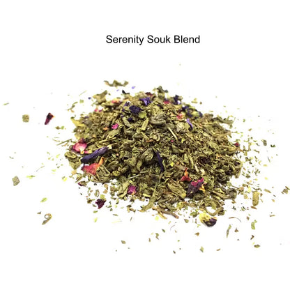 A small pile of loose herbal tea blend labeled 'Serenity Souk Blend' on a plain white background. The blend is primarily green with visible dried flower petals in pink, red, and purple tones.