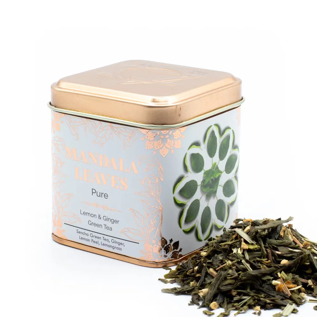 A square metal tin of Mandala Leaves Pure Lemon & Ginger Green Tea with a rose gold lid and a white label featuring a green mandala design, shown next to a small pile of loose tea leaves on a white background.
