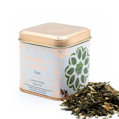 A square metal tin of Mandala Leaves Pure Lemon & Ginger Green Tea with a rose gold lid and a white label featuring a green mandala design, shown next to a small pile of loose tea leaves on a white background.