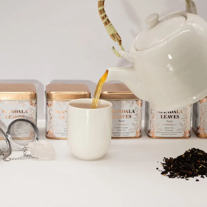White teapot pouring tea into a white mug with tea packaging in the background.